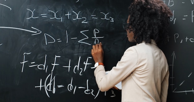 Vista posterior de la profesora joven afroamericana en la escuela escribiendo fórmulas y leyes matemáticas en la pizarra. Concepto de escuela. Profesora de gafas explicando las leyes de la física. Posterior.