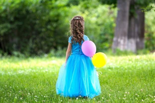 Vista posterior del retrato de cuerpo entero de una bonita niña rubia de pelo largo con un vestido largo azul con globos de colores de pie en el campo floreciente en árboles verdes borrosos