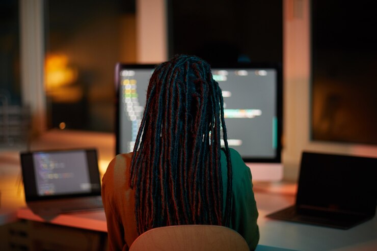 Foto vista posterior retrato de desarrollador de ti femenino escribiendo código en varias pantallas de computadora mientras trabaja hasta tarde en la oficina oscura, espacio de copia