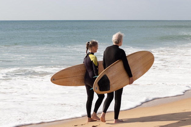 Vista posterior de los surfistas de edad caminando por la orilla del mar. Hombre y mujer deportivos con trajes de neopreno sosteniendo tablas de surf paseando por la playa de arena disfrutando de las vistas de las olas del mar azul. Estilo de vida saludable del concepto de personas mayores