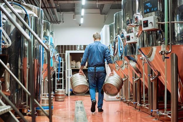 Vista posterior del trabajador masculino de la planta de producción de cerveza contemporánea que lleva dos cisternas mientras se mueve a lo largo de filas de enormes tanques de acero