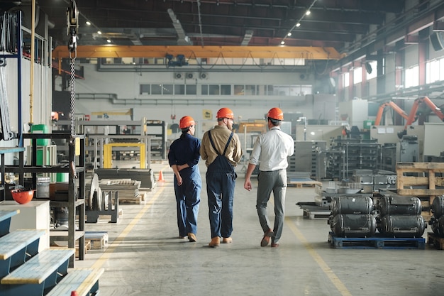 Foto vista posterior de tres jóvenes trabajadores contemporáneos de la planta industrial caminando a lo largo de una gran fábrica y comunicándose