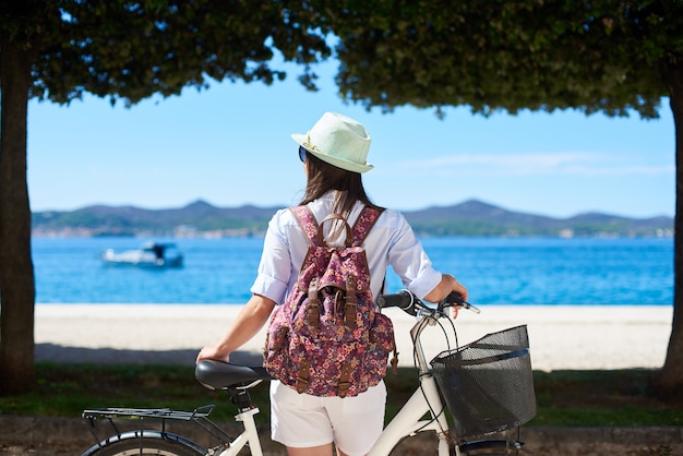 Vista posterior turista femenina de pie con bicicleta en la acera cerca de la costa bajo árboles y montañas