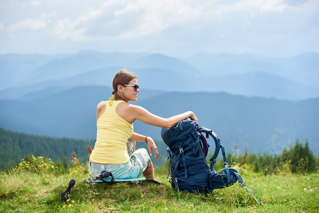 Vista posterior del turista mujer atractiva sentada en la colina cubierta de hierba con mochila