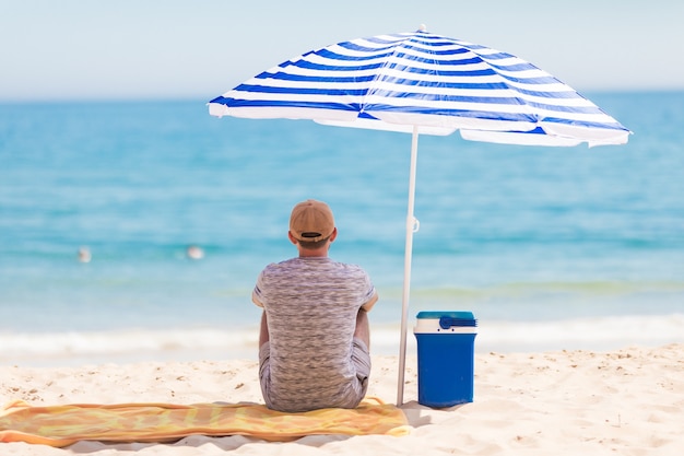 Vista posterior del turista sentado en la playa bajo la sombrilla cerca del enfriador con bebidas frías