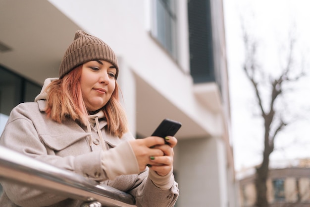 Vista de primer plano en ángulo bajo de una mujer con sobrepeso en un sombrero de otoño escribiendo un mensaje en línea, navegando en las redes sociales usando un teléfono móvil parado en la calle de la ciudad en el fondo del edificio de oficinas en un día nublado