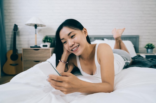 vista de primer plano la joven asiática acostada sobre la cara apoyada en el vientre en el dormitorio está cargando fotos en el sitio web de las redes sociales. atractiva chica taiwanesa está holgazaneando phubbing en la cama.