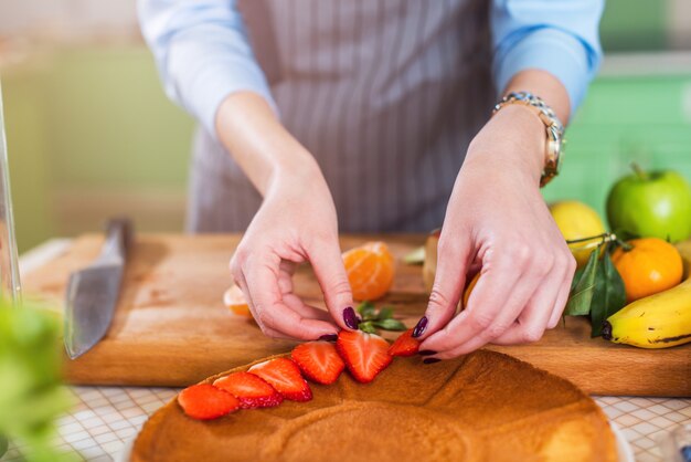 Vista de primer plano de manos femeninas poner rodajas de fresa en una capa de pastel. Mujer haciendo pastel de bayas en la cocina