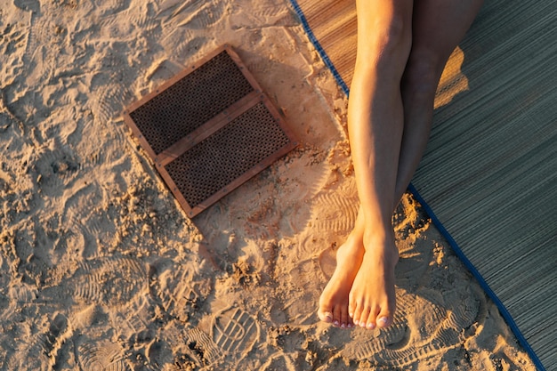 Vista de primer plano de una mujer irreconocible sentada en una alfombra de yoga con una tabla Sadhu en una playa de arena en la mañana soleada de verano