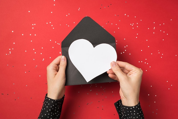 Vista en primera persona arriba de la foto de manos femeninas sosteniendo un sobre negro con una tarjeta blanca como corazón dentro aislado en el fondo rojo con confeti