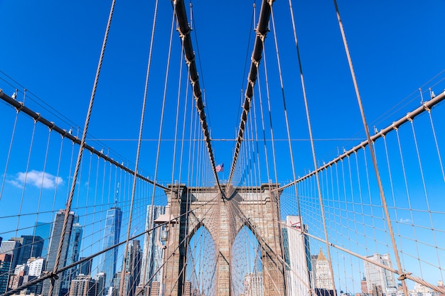 Vista del Puente de Brooklyn con tirantes diagonales y tirantes verticales. En el centro de la foto está la bandera estadounidense ondeando al viento.