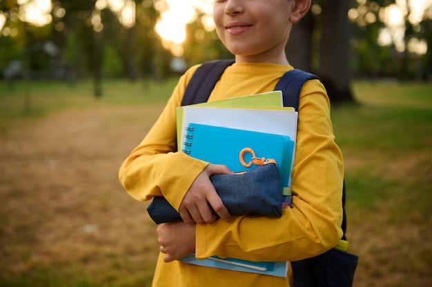 Vista recortada de un adorable niño de escuela sonriente con útiles escolares y libros de trabajo en las manos posando para la cámara en el parque de la ciudad al atardecer