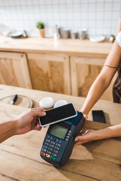 Vista recortada del cajero que sostiene la terminal mientras el hombre paga con el teléfono inteligente en la cafetería