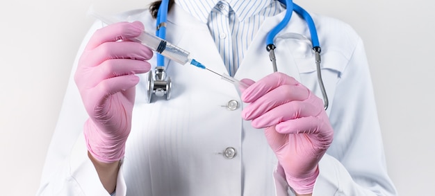 Vista recortada da médica em um jaleco branco e luvas estéreis rosa segurando a seringa com injeção.