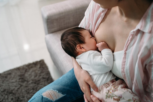 vista recortada de cima, o pequeno bebê recém-nascido está aninhado em sua mãe enquanto chupa o leite materno na sala de estar em casa.