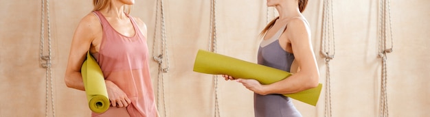 Foto vista recortada de duas mulheres vestindo roupas esportivas em pé na academia segurando o tapete de ioga, prontas para iniciar a preparação física
