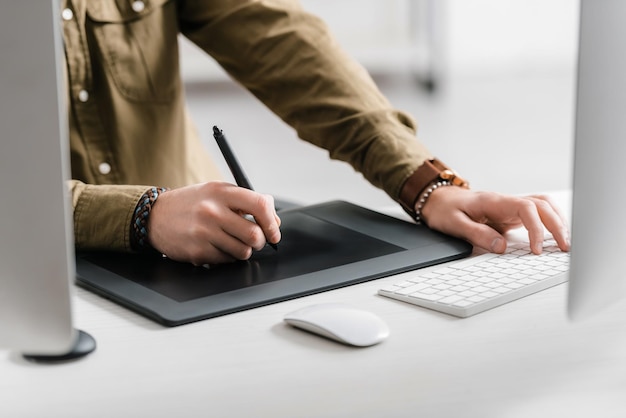 Vista recortada del diseñador que trabaja con tableta gráfica y teclado de computadora en la mesa