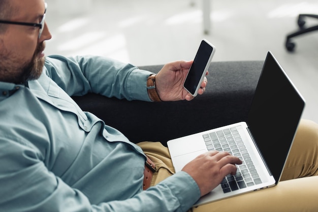 Foto vista recortada del hombre de negocios en camisa con computadora portátil y teléfono inteligente en la oficina