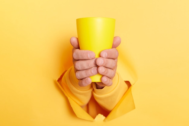 Vista recortada del hombre que sostiene el café en la taza con ambas manos a través del agujero en la pared de papel aislado sobre fondo amarillo