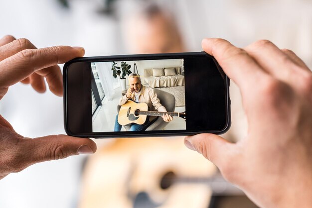 Foto vista recortada de un hombre tomando una foto de un padre mayor sosteniendo una guitarra acústica en casa