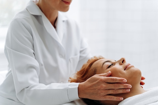 Vista recortada de las manos del masajista masajeando la cara de la encantadora dama en el salón de spa. Mujer tendida en el sofá y recibiendo procedimientos de spa