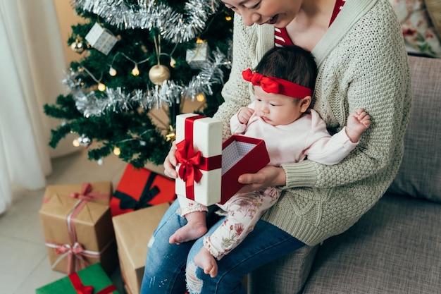 vista recortada mujer asiática está jugando con su hijo pequeño el día del boxeo. padre milenario sonriente mostrando el interior de la caja de regalo a su bebé curioso. momentos reales