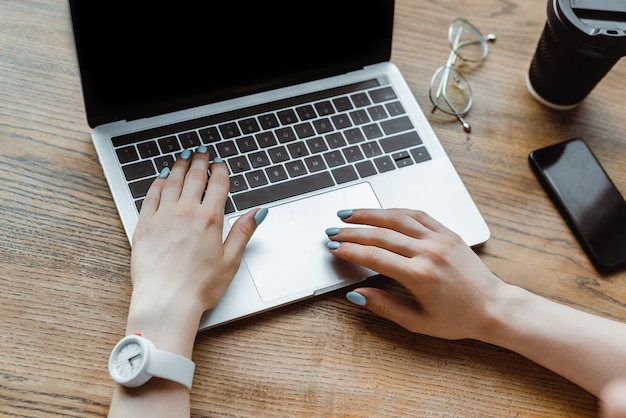 Vista recortada de mujer escribiendo en el teclado del ordenador portátil cerca de teléfono inteligente y café para ir a la mesa de madera en la cafetería