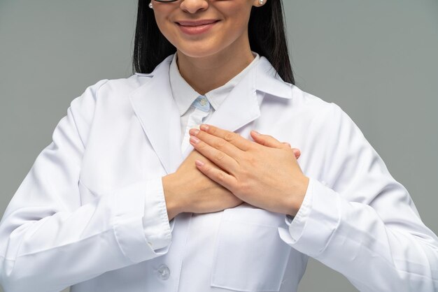 Vista recortada de la mujer morena sonriendo a la cámara mientras sostiene las manos en su pecho Retrato de una hermosa joven doctora en una chaqueta médica blanca aislada en un fondo gris