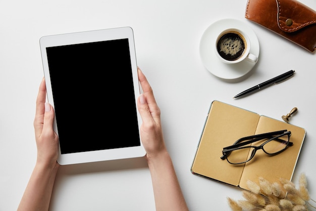 Vista recortada de la mujer que sostiene la tableta digital en las manos cerca de los vasos de café del bloc de notas y el estuche en