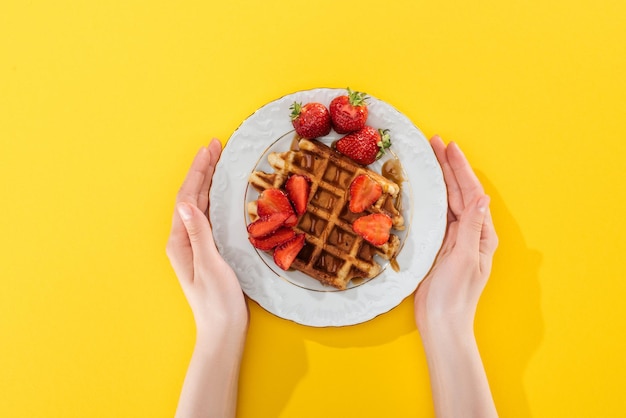 Vista recortada de mujer sosteniendo plato con waffle y fresas en amarillo