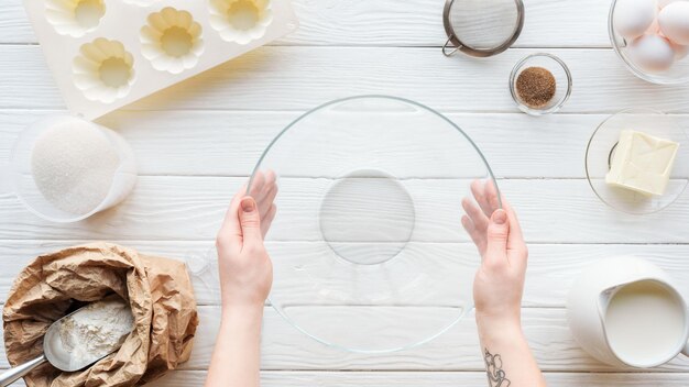 Vista recortada de la mujer sosteniendo un tazón de vidrio en la mesa con utensilios de cocina e ingredientes