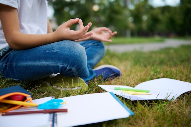 Vista recortada del niño de la escuela sentado en posición de loto sobre la hierba verde del parque de la ciudad y meditando. Libros de trabajo útiles escolares tirado en el césped. Conceptos de concentración, recreación, atención plena