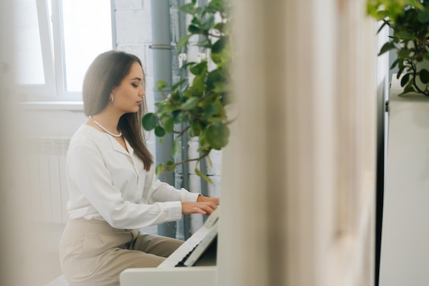 Vista remota de una talentosa joven pianista tocando un piano clásico blanco en el aula durante la lección Lección de piano de una estudiante de música enfocada mirando notas musicales en la sala de estar