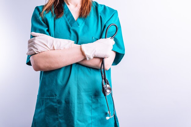 Vista selectiva de los brazos cruzados de la doctora del hospital sosteniendo un estetoscopio con guantes sobre fondo blanco.