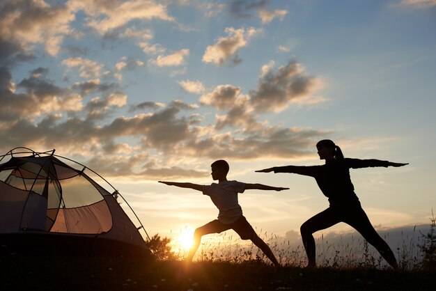 Vista de la silueta de Yoga Warrior pose ejercicio con pareja cerca de la tienda al amanecer