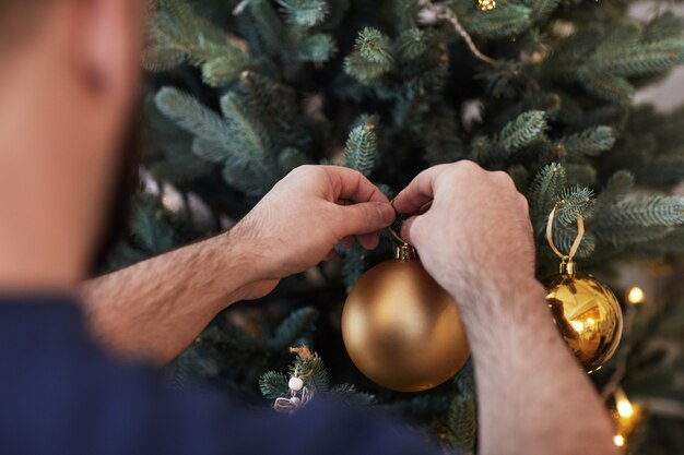 Vista sobre el hombro del hombre que cuelga la bola de Navidad dorada en el árbol mientras se prepara para la celebración