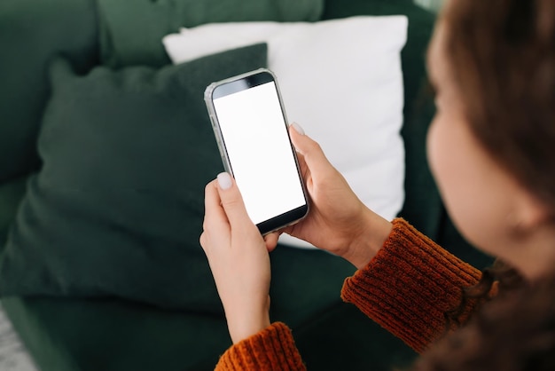 Vista sobre o ombro de uma mulher segurando um smartphone com uma tela de celular simulada branca em mãos usando