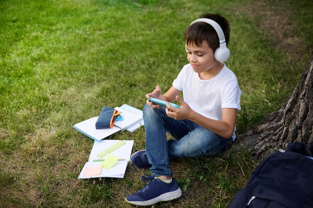 Vista superior. Adorable y guapo colegial de primaria con auriculares inalámbricos, centrado en jugar con el teléfono inteligente, descansando sobre la hierba verde del parque urbano, útiles escolares acostado junto a él