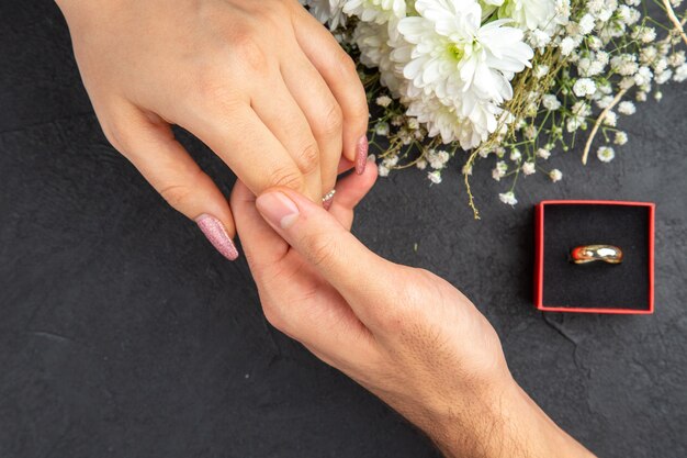 Vista superior alcançando as mãos, mão masculina segurando o buquê de flores do anel de noivado feminino em fundo escuro