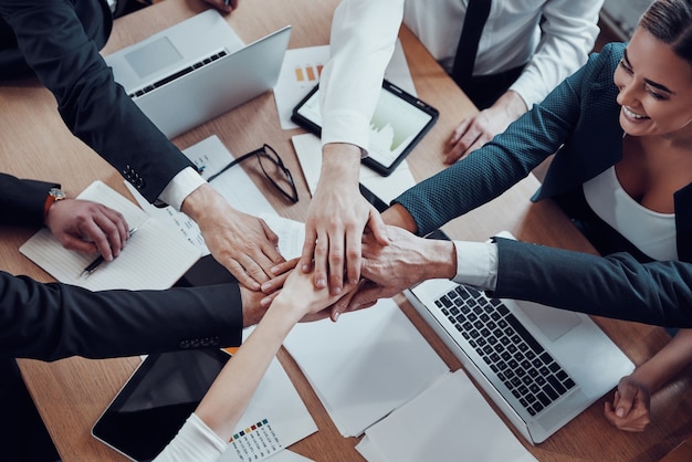 Vista superior del alegre equipo de negocios manteniendo las manos juntas como símbolo de unidad mientras trabajan juntos en la oficina moderna