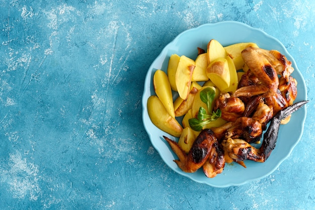 Vista superior de alitas de pollo en adobo de salsa de soja y rodajas de papas al horno en placa azul sobre fondo azul con espacio de copia