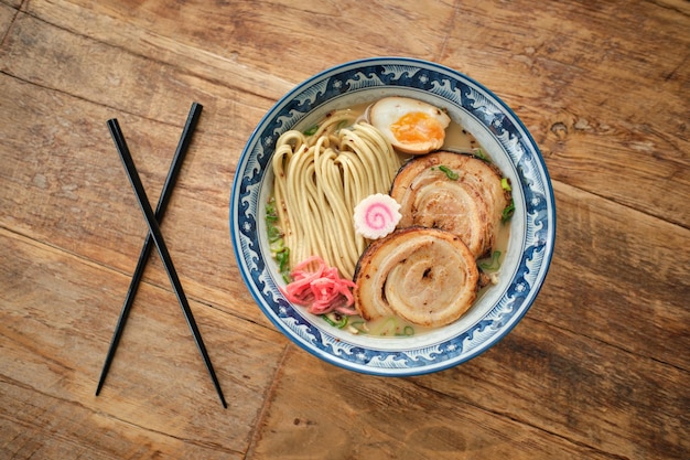 Vista superior de la apetitosa sopa de ramen con fideos de huevo cocidos y carne servida en un tazón de cerámica colocado cerca de palillos en una mesa de madera