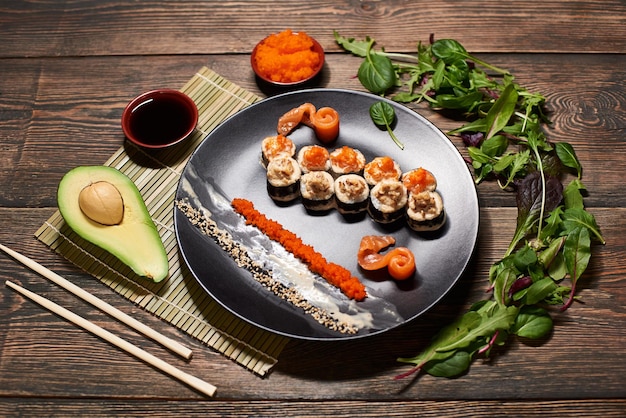 Vista superior de apetitosos rollos de sushi japoneses en un gran plato negro que sirve con rodajas de pescado, salsas, caviar, ensalada de aguacate