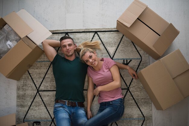 Foto vista superior de una atractiva pareja joven moviéndose, tomándose de la mano, mirando a la cámara y sonriendo mientras yacía entre cajas de cartón