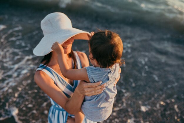 Vista superior atraente da jovem mãe brincando com a filha com chapéu branco no fundo do mar Férias em família Viagem Jogo feminino com criança alegre Mulher caucasiana com bebê ao pôr do sol do oceano