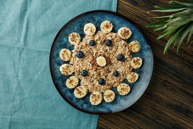 Vista superior de avena con nuez de endrino de plátano y sésamo en plato sobre tela azul con hojas de piña sobre fondo de madera