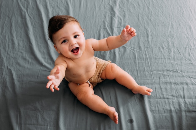 Vista superior del bebé sonriente sentado en la cama en la habitación con pantalones cortos