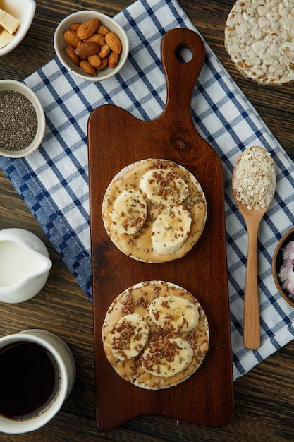 Vista superior de bocadillos de pan crujiente de mantequilla de maní de plátano en una tabla de cortar con pan crujiente de leche de almendras y café de avena sobre tela escocesa sobre fondo de madera