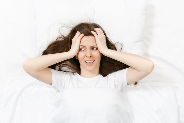 Vista superior de la cabeza de la mujer joven morena cansada acostada en la cama con sábana blanca, almohada, manta. Mujer sorprendida cubre las orejas con la mano, pasando tiempo en la habitación