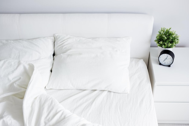 Foto vista superior de la cama vacía sin hacer con una manta blanca y una mesita de noche con dos almohadas con despertador y una maceta en casa